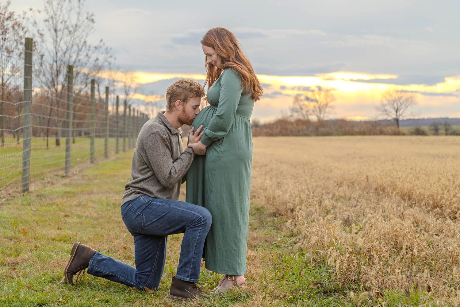 Man kneeling before pregnant woman in green dress at sunset in a field, romantic maternity photo session outdoors.