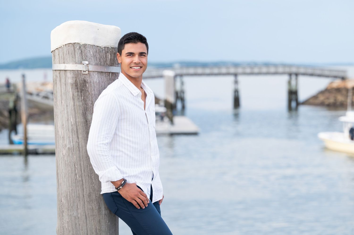 A person in white clothing walks along a coastal pier with a lighthouse and boats visible in the distance.