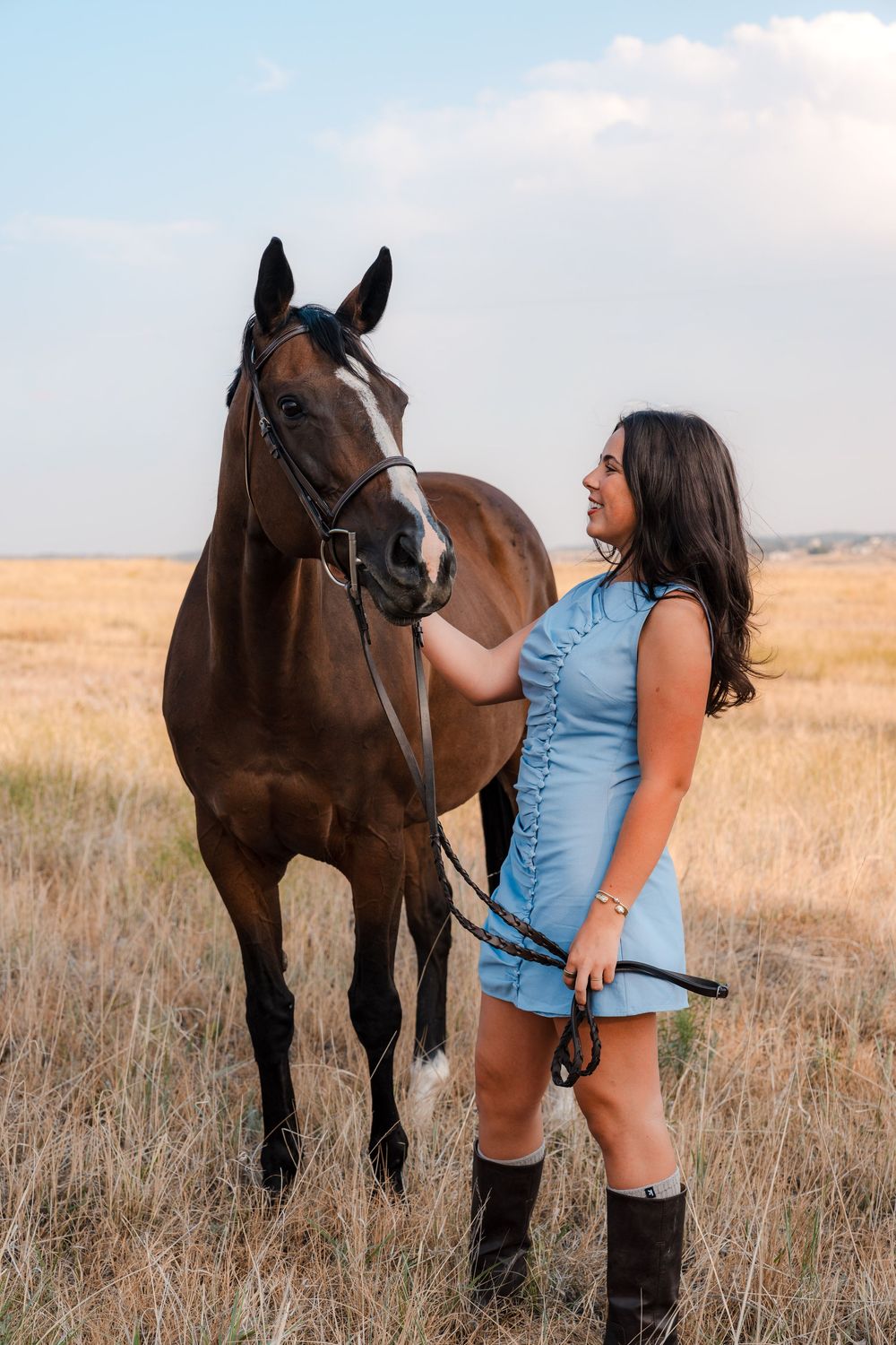 Young woman in a light blue dress and tall black boots stands beside a brown bridled horse in a golden grassy field under a soft sky.