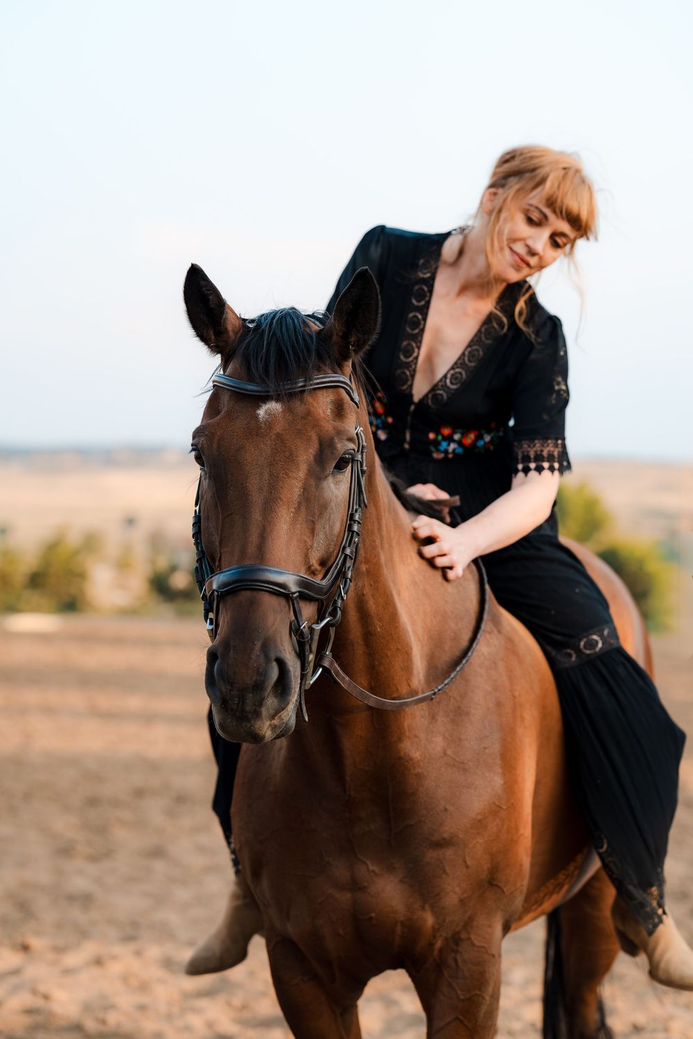 Woman in a black embroidered blouse and long skirt sits on a brown bridled horse in a sandy field at sunset.