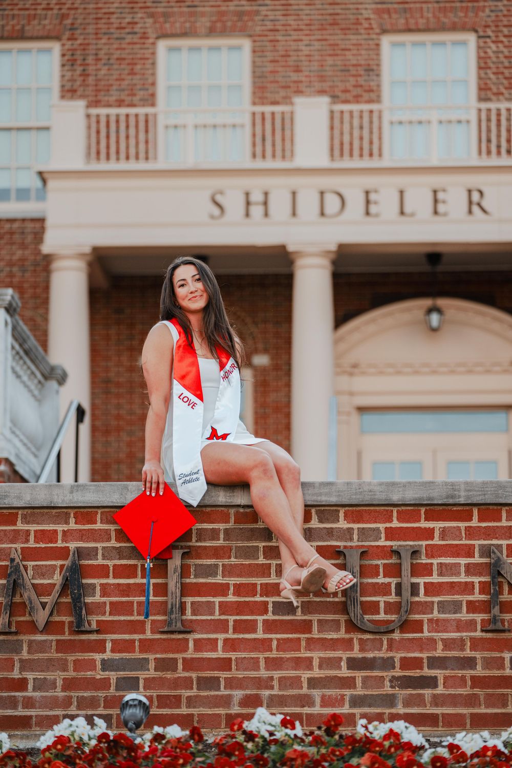Female graduate sitting on brick wall outside Shideler Hall holding red cap.