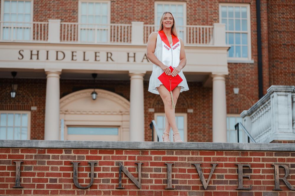 Blonde graduate in red and white dress standing on brick wall outside Shideler Hall university building.
