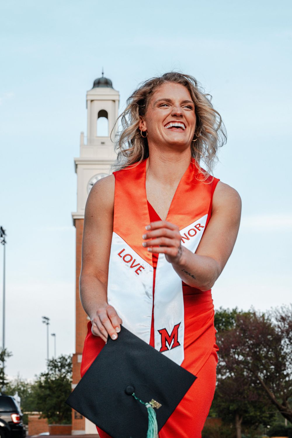 Smiling female graduate in red gown holding cap near university bell tower.