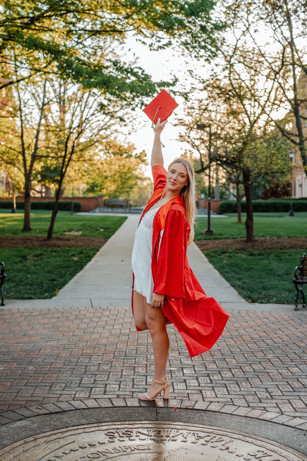 Female graduate in red gown raising cap triumphantly on brick campus pathway.
