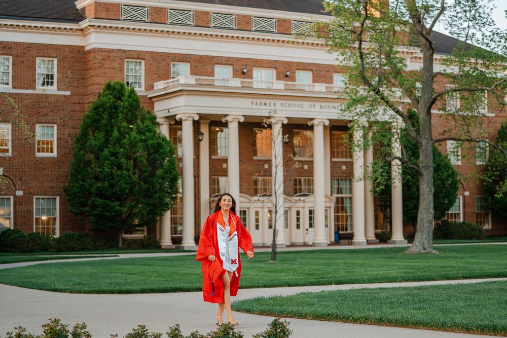Female graduate in red gown walking outside a columned university building.