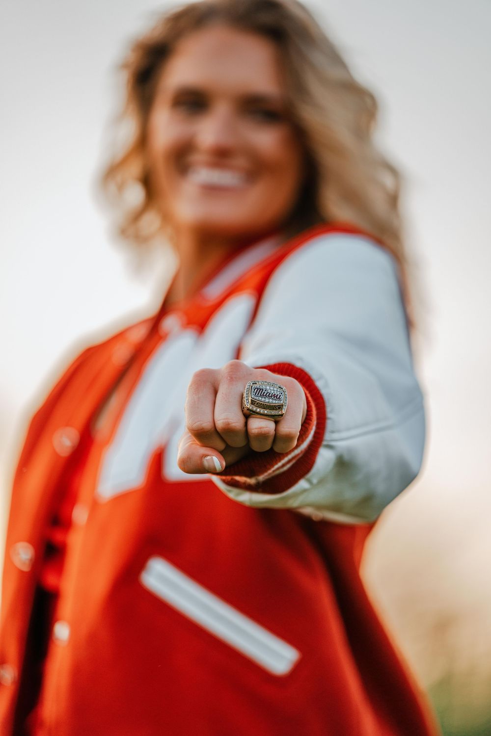Female graduate in red varsity jacket showing championship ring toward camera.