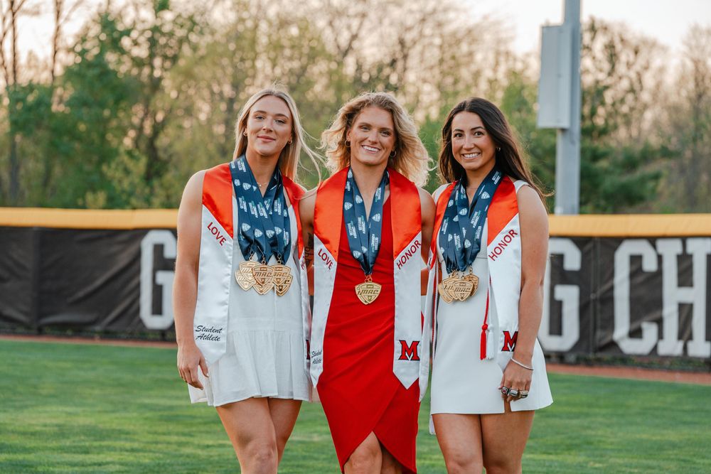 Three female graduates wearing medals standing on baseball field with Graduating Champions banner.