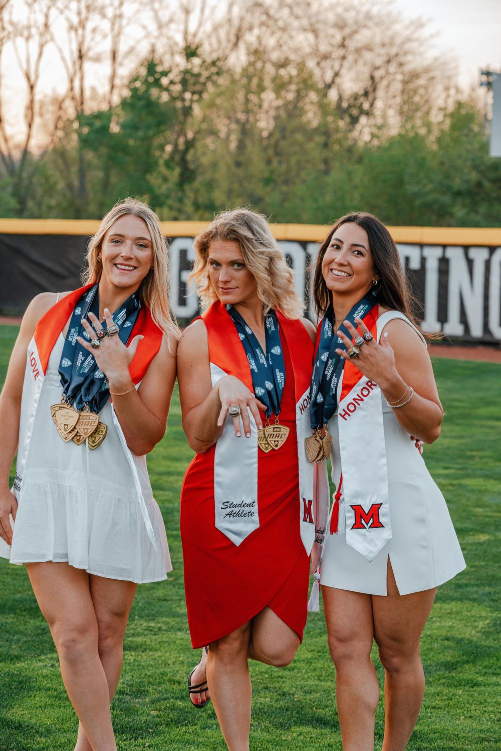 Three female graduates in red and white dresses display rings and medals on a sports field.