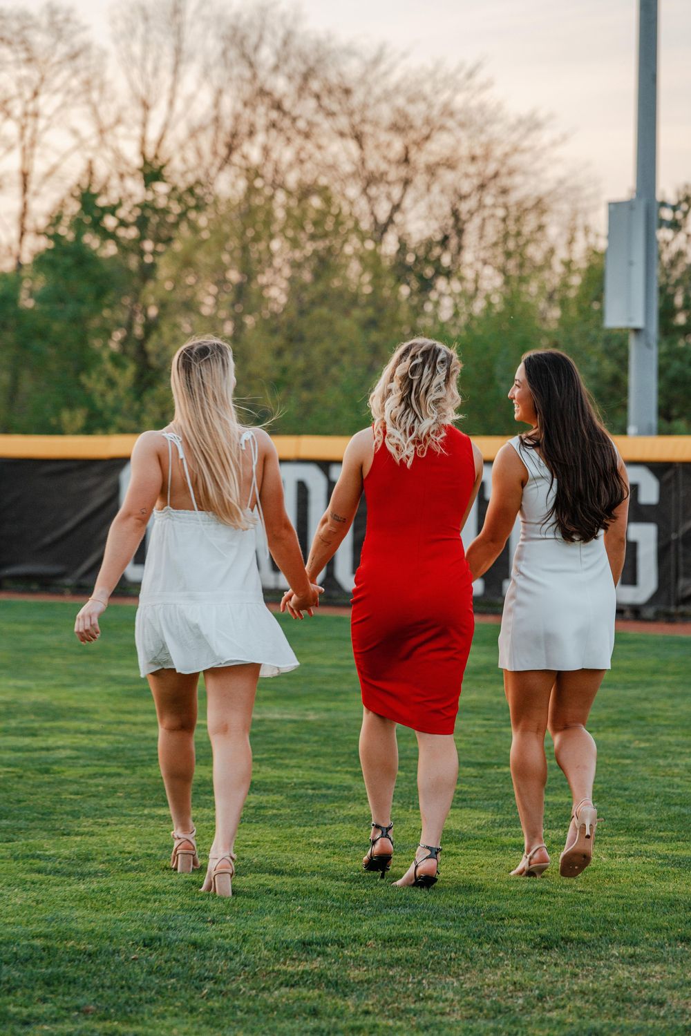 Three female graduates in red and white dresses walk hand-in-hand across a green sports field.