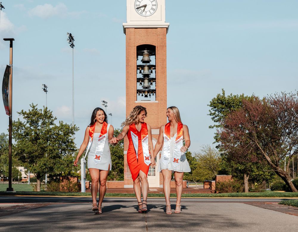 Three female graduates in red and white stoles walk past campus clock tower.