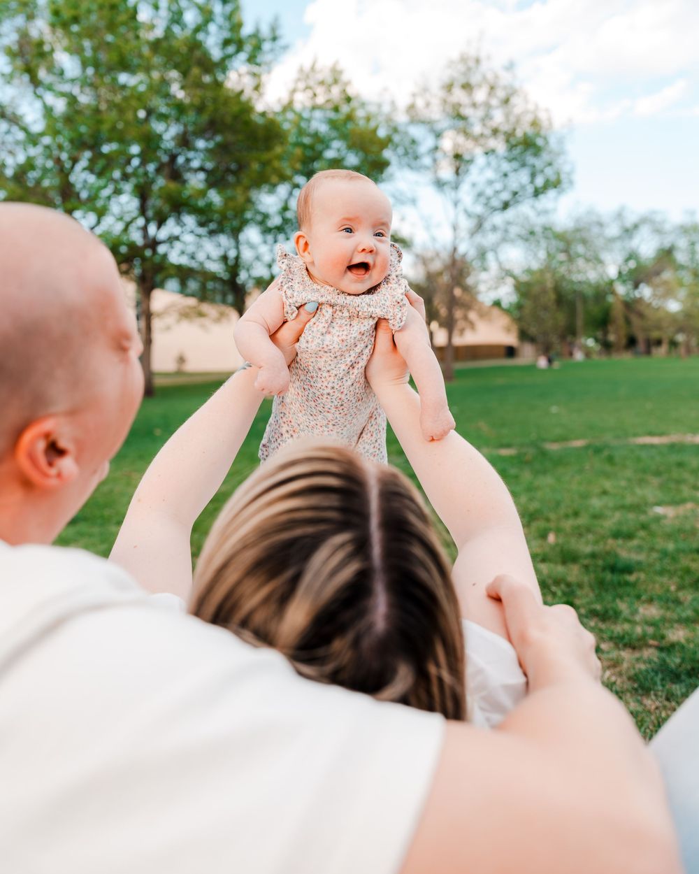 Parents joyfully lifting their laughing baby girl in a sunny green park.