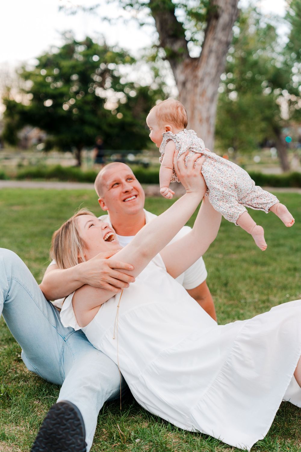Happy family with parents sitting on grass in park, joyfully lifting smiling baby girl in floral outfit on sunny day.