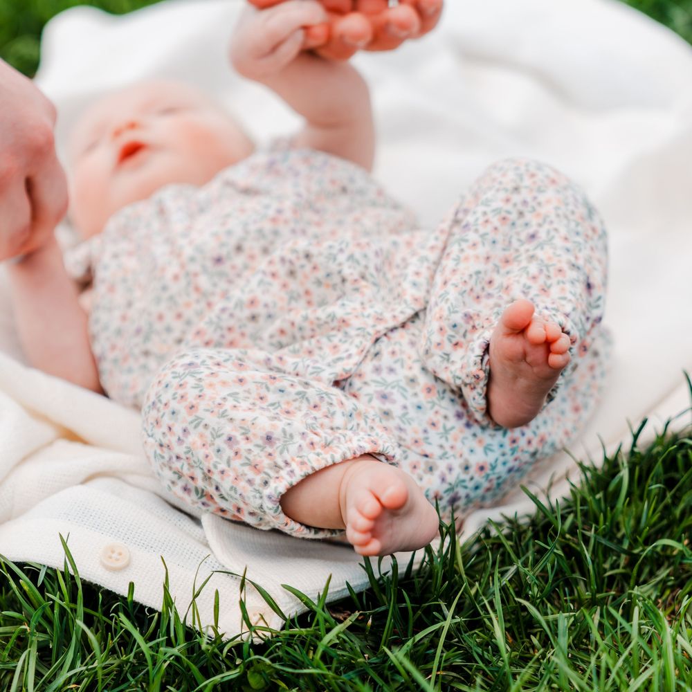 Baby in floral romper lying on white blanket on green grass, tiny feet and toes visible in outdoor setting.