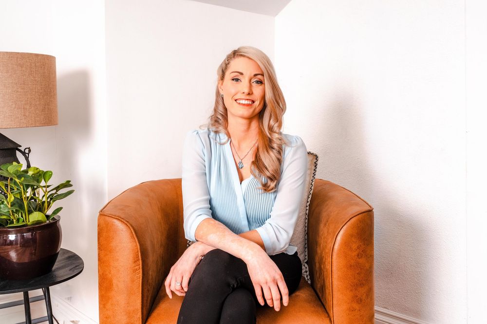 Smiling blonde woman in light blue blouse sitting in brown leather armchair in modern interior.