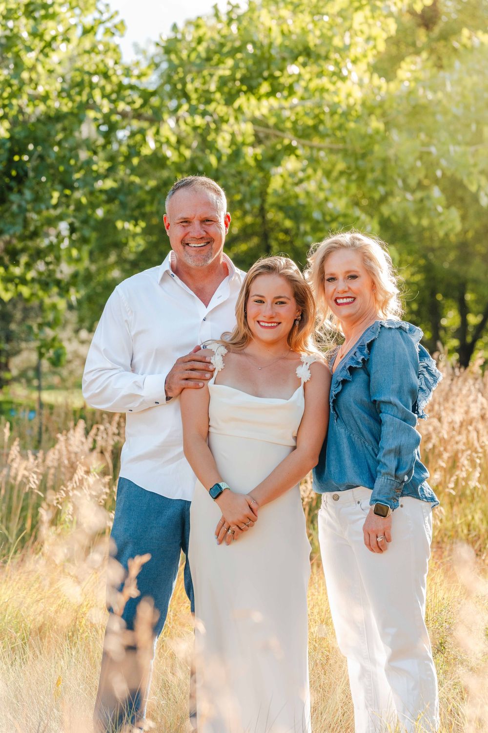 Smiling family of three poses together in a sunny field with lush green trees in the background.