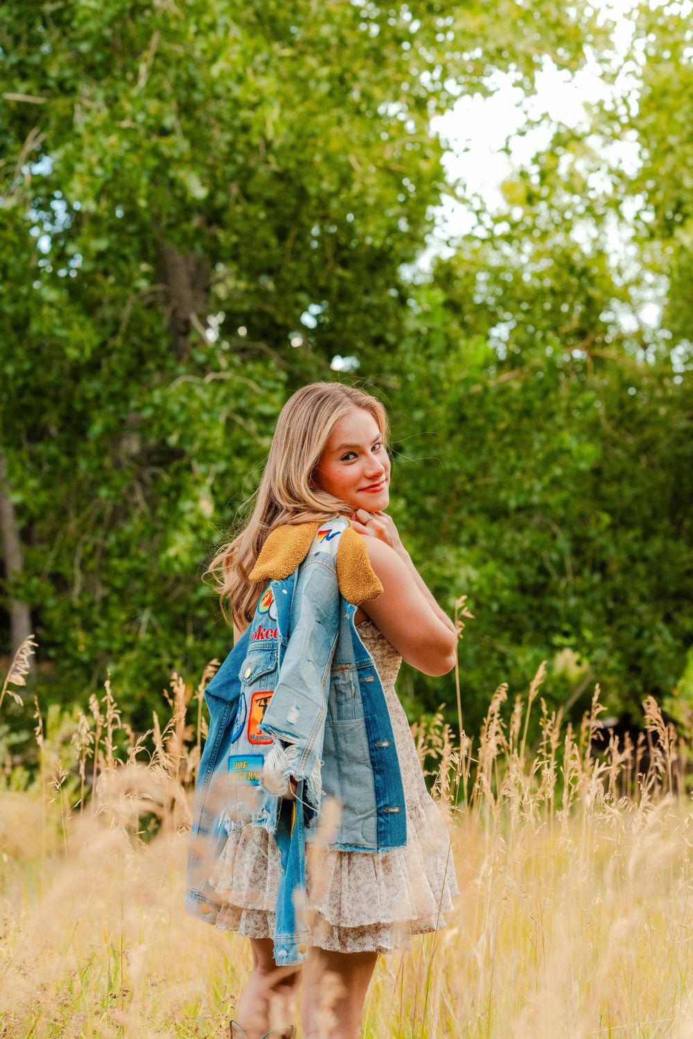 Smiling blonde woman in lace dress and denim jacket posing in sunlit meadow with green trees.