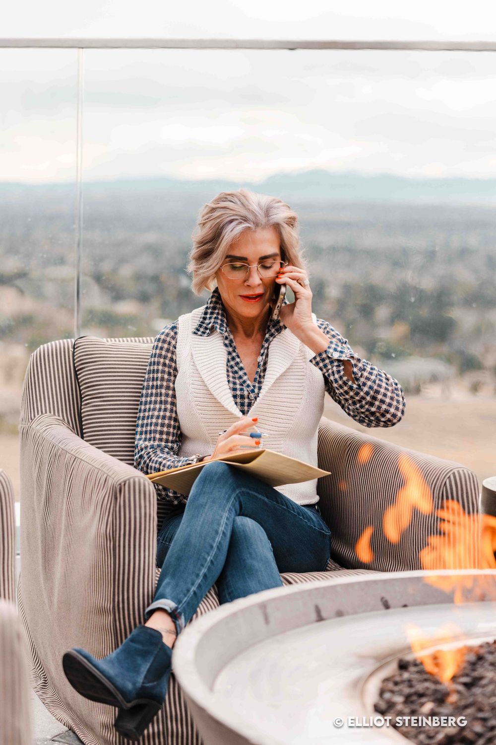 Mature woman multitasking on phone and notebook beside outdoor fire pit with mountain view backdrop.