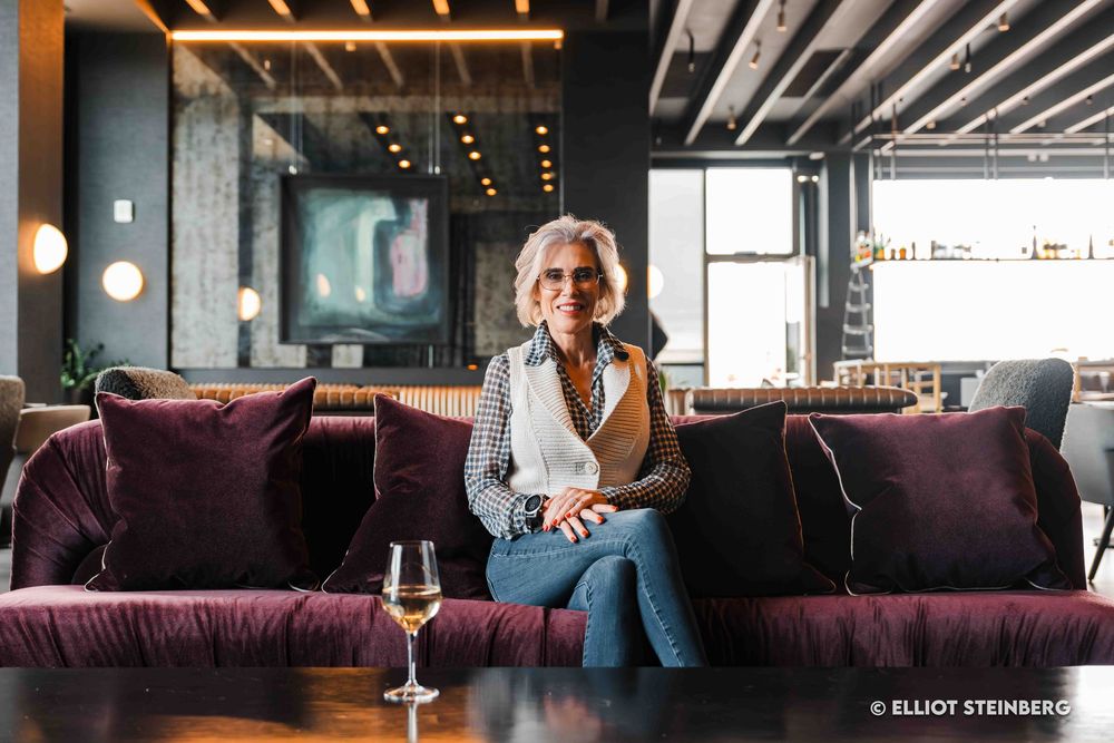 Elegant older woman sitting on purple velvet sofa with wine glass in modern upscale bar interior.