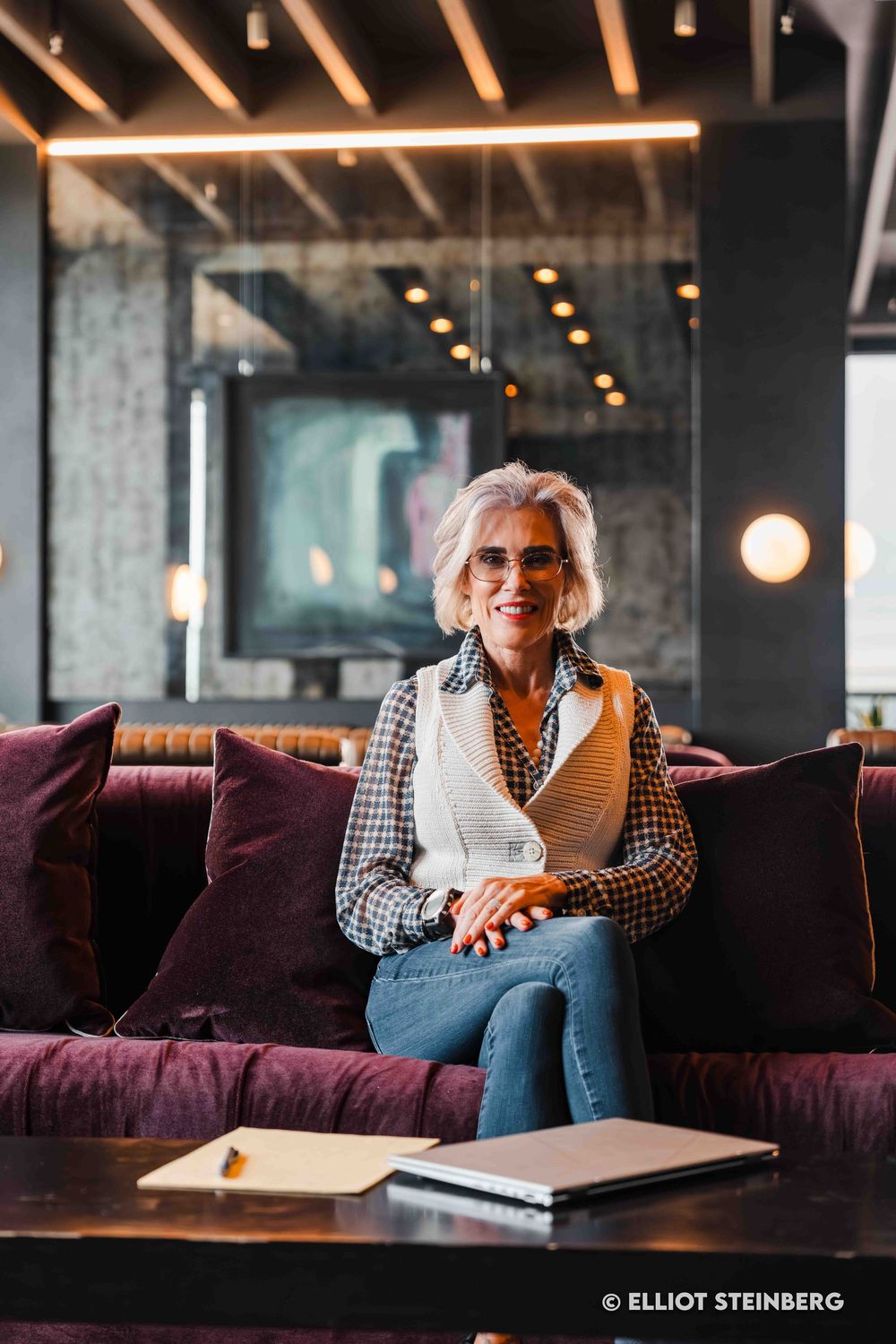 Stylish senior woman with gray hair and glasses sitting on purple velvet couch in modern lounge.
