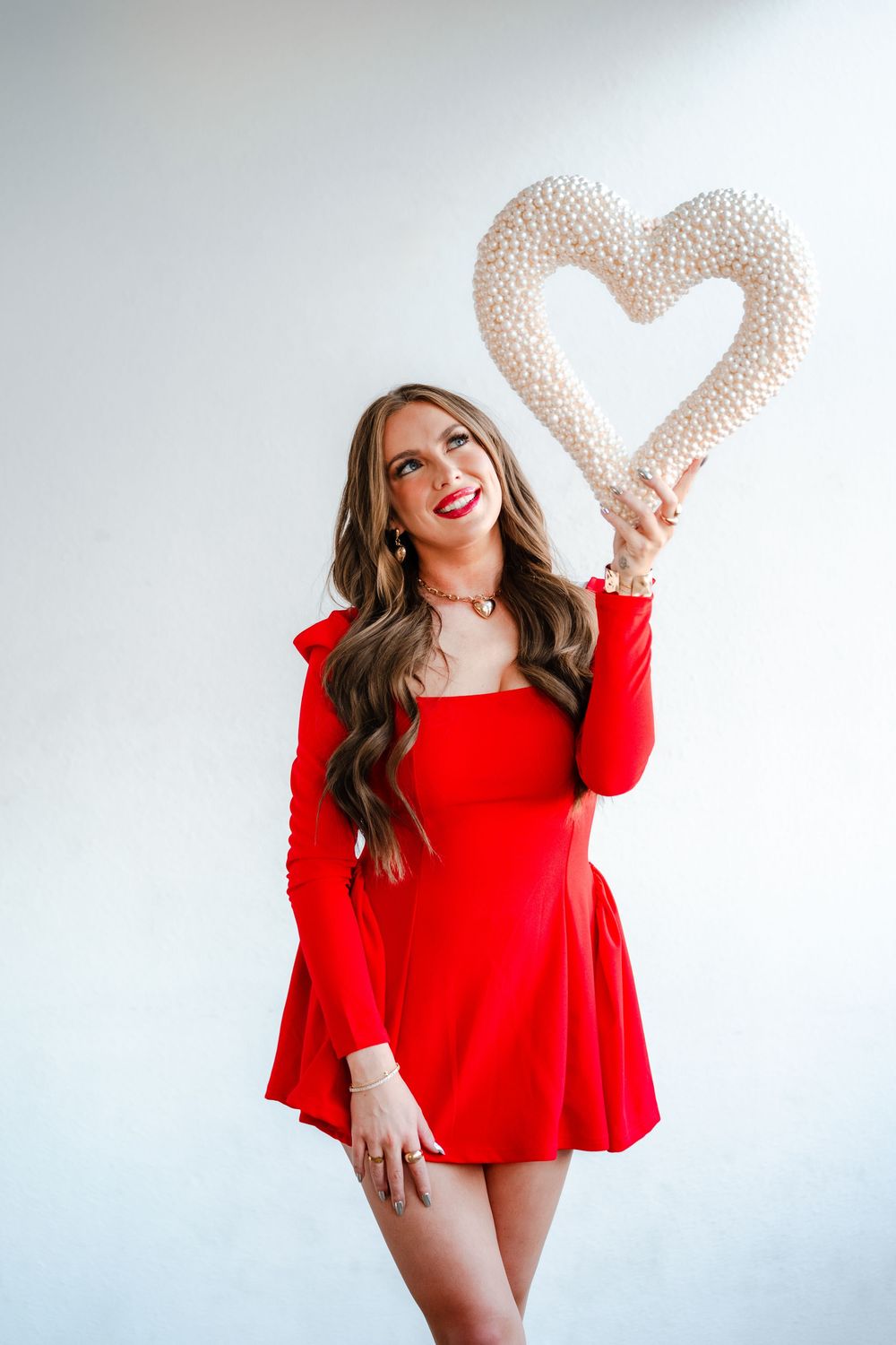 Brunette woman in red long-sleeve mini dress holding pearl heart decoration overhead.