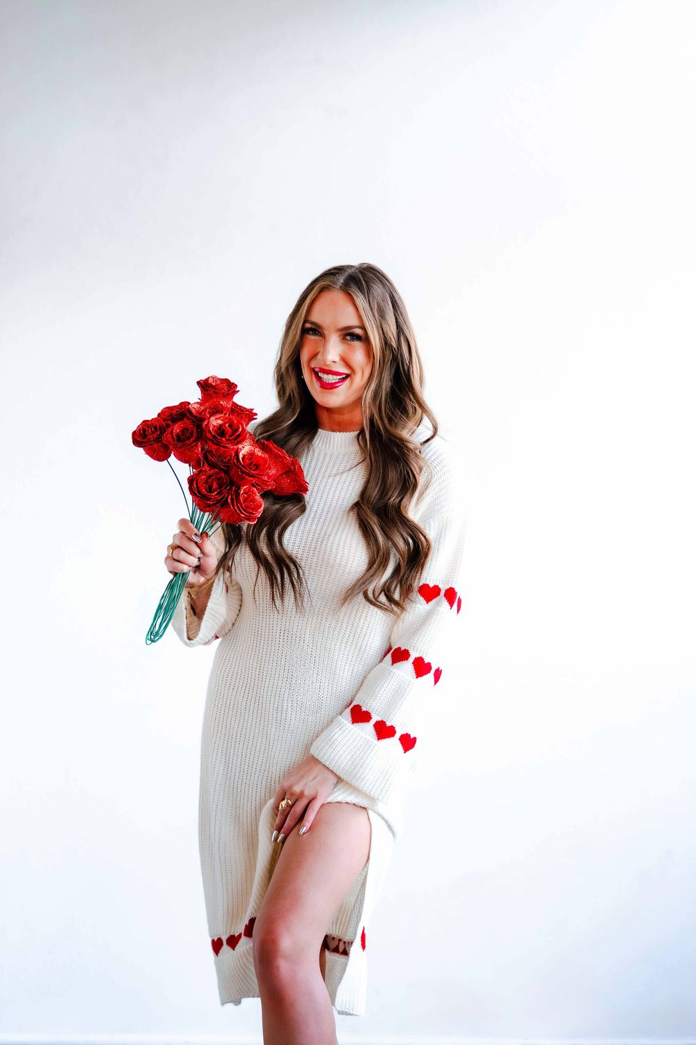 Brunette woman in white heart-print sweater dress holding bouquet of red roses smiling.