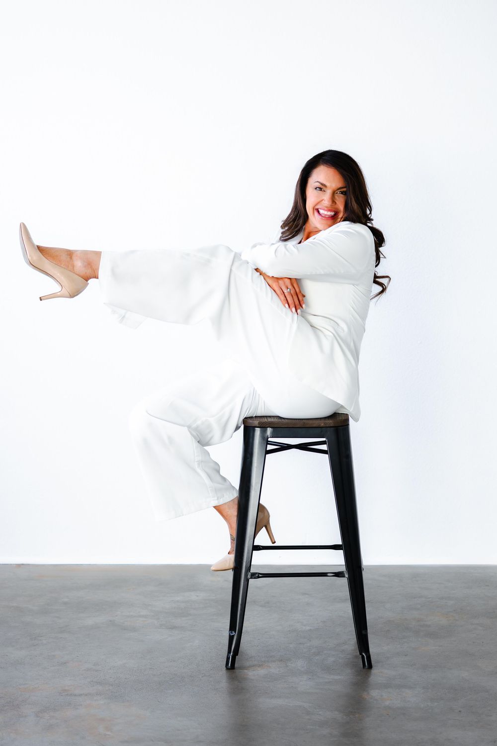 Woman in white outfit sitting on black stool with leg extended, smiling confidently against white background.