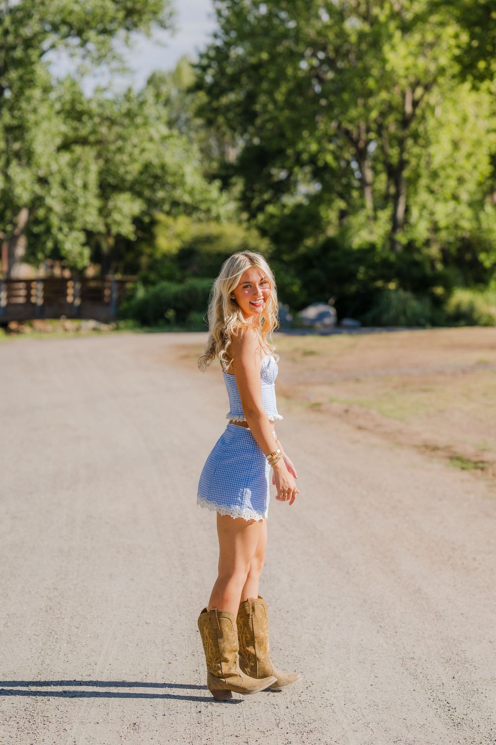 Blonde woman in denim mini skirt, white top, and cowboy boots standing on a gravel road surrounded by trees.