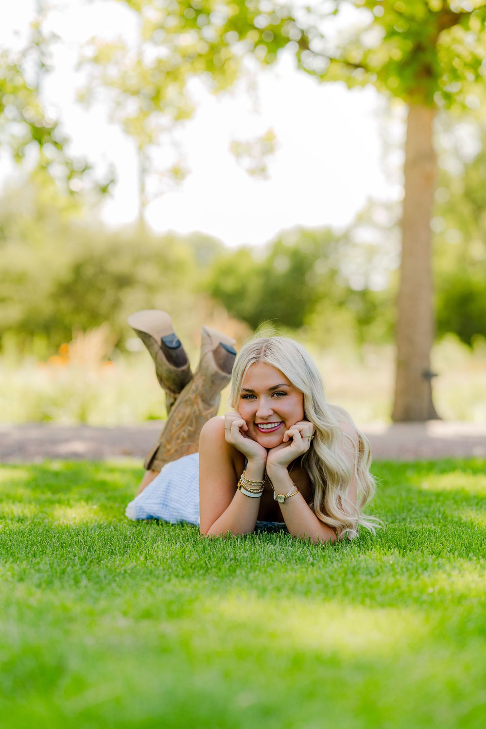 Smiling blonde woman lying on green grass in a white dress and cowboy boots in a sunny park setting.