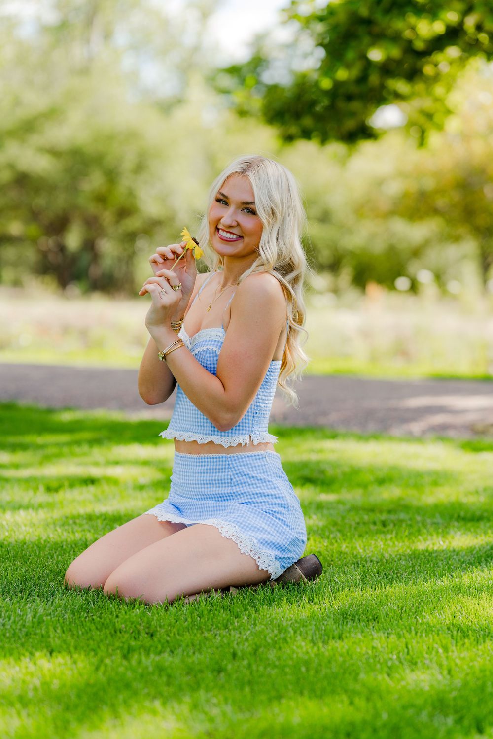 Smiling blonde woman in blue gingham crop top and skirt kneeling on green grass in a sunny park holding a yellow flower.