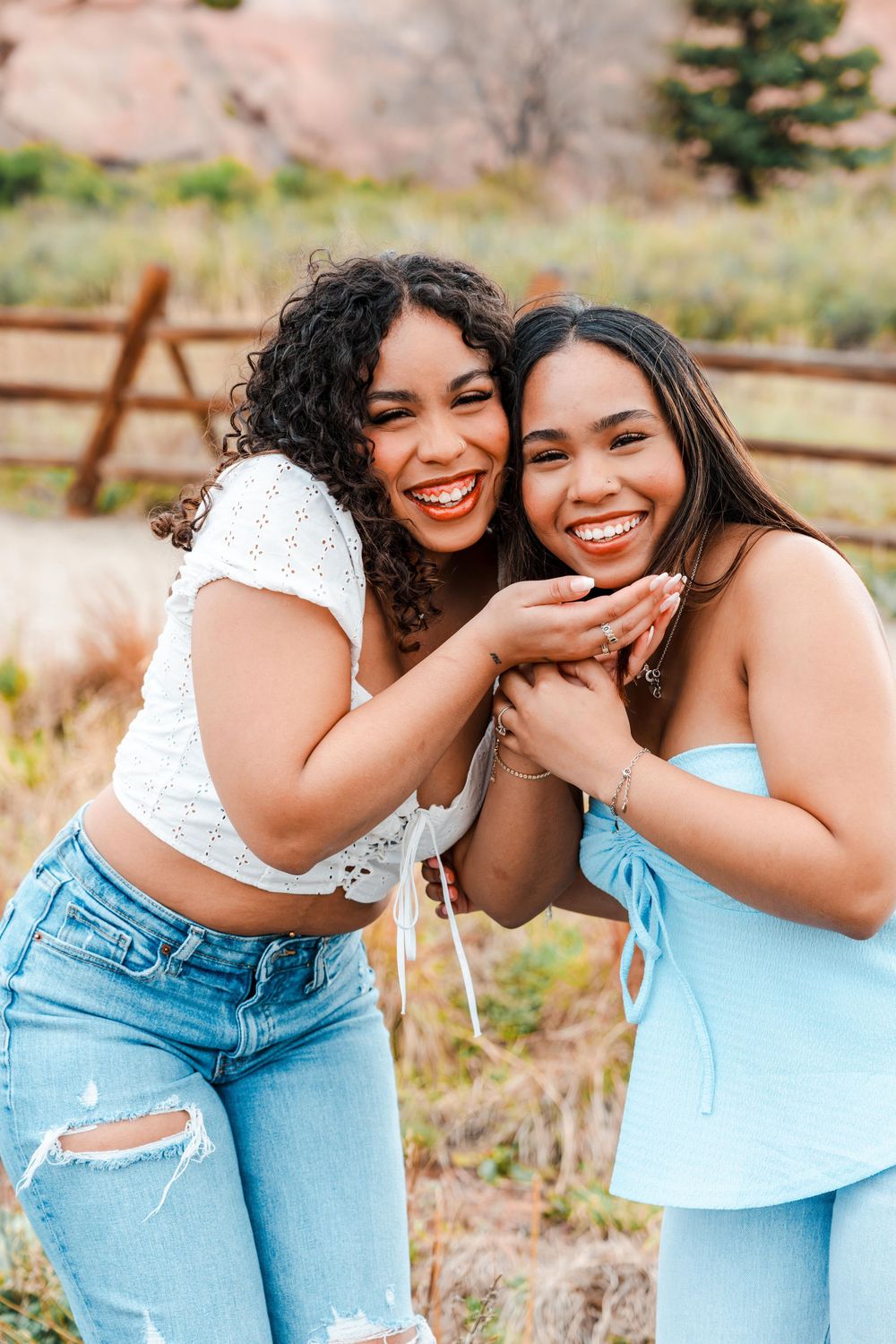 Two smiling young women in blue and white outfits embrace outdoors near a wooden fence on a sunny day.