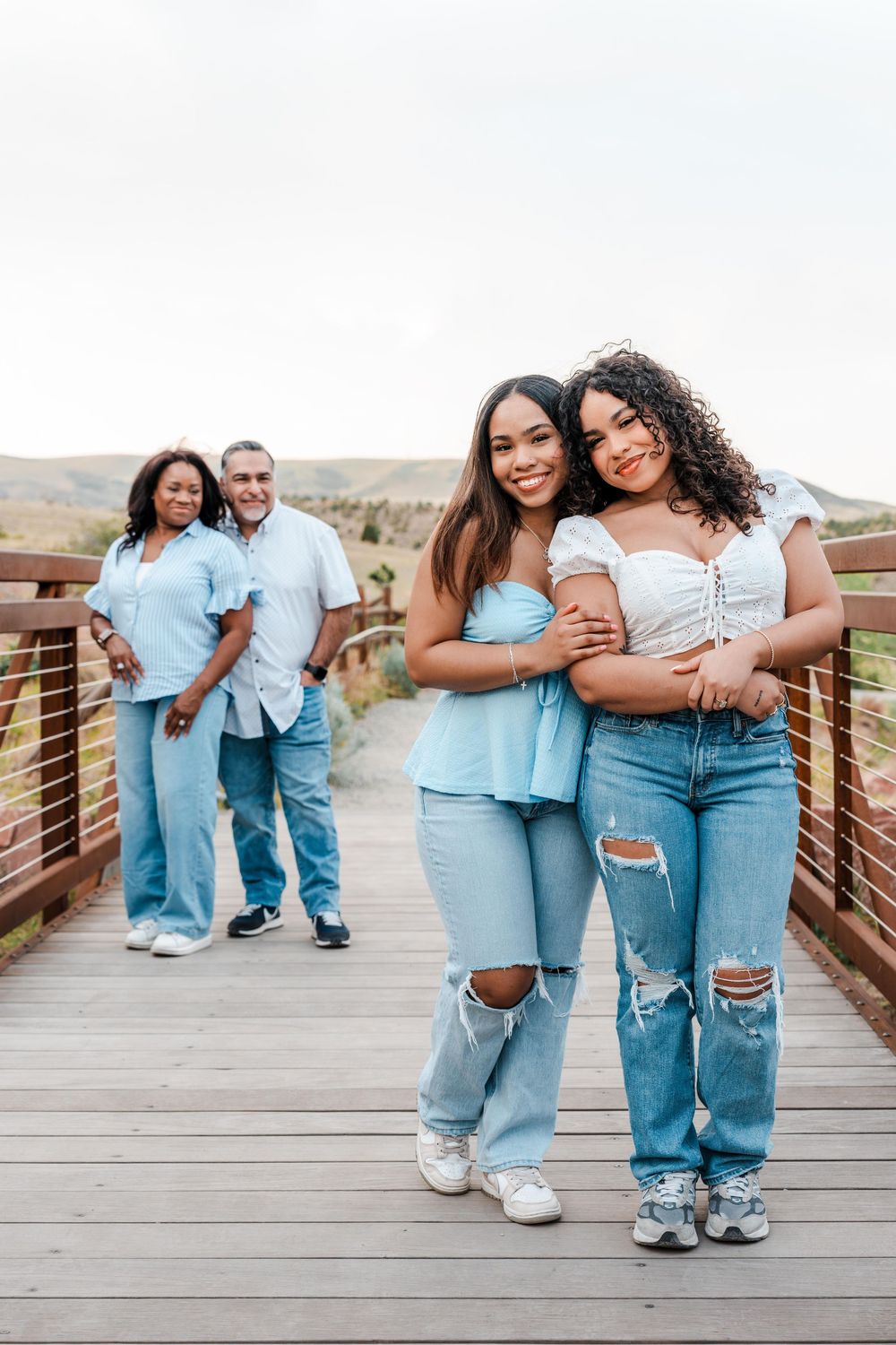 Two young women hugging on a wooden bridge outdoors, with a couple standing behind them, all wearing blue and white outfits.