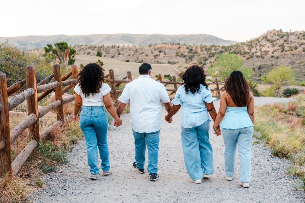 Family of four holding hands walking down a dirt path surrounded by scenic desert hills and a wooden fence.