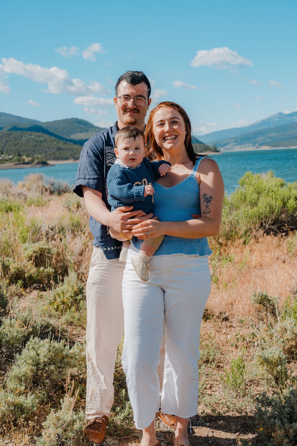 A young family with a baby poses outdoors near a mountain lake under a blue sky.