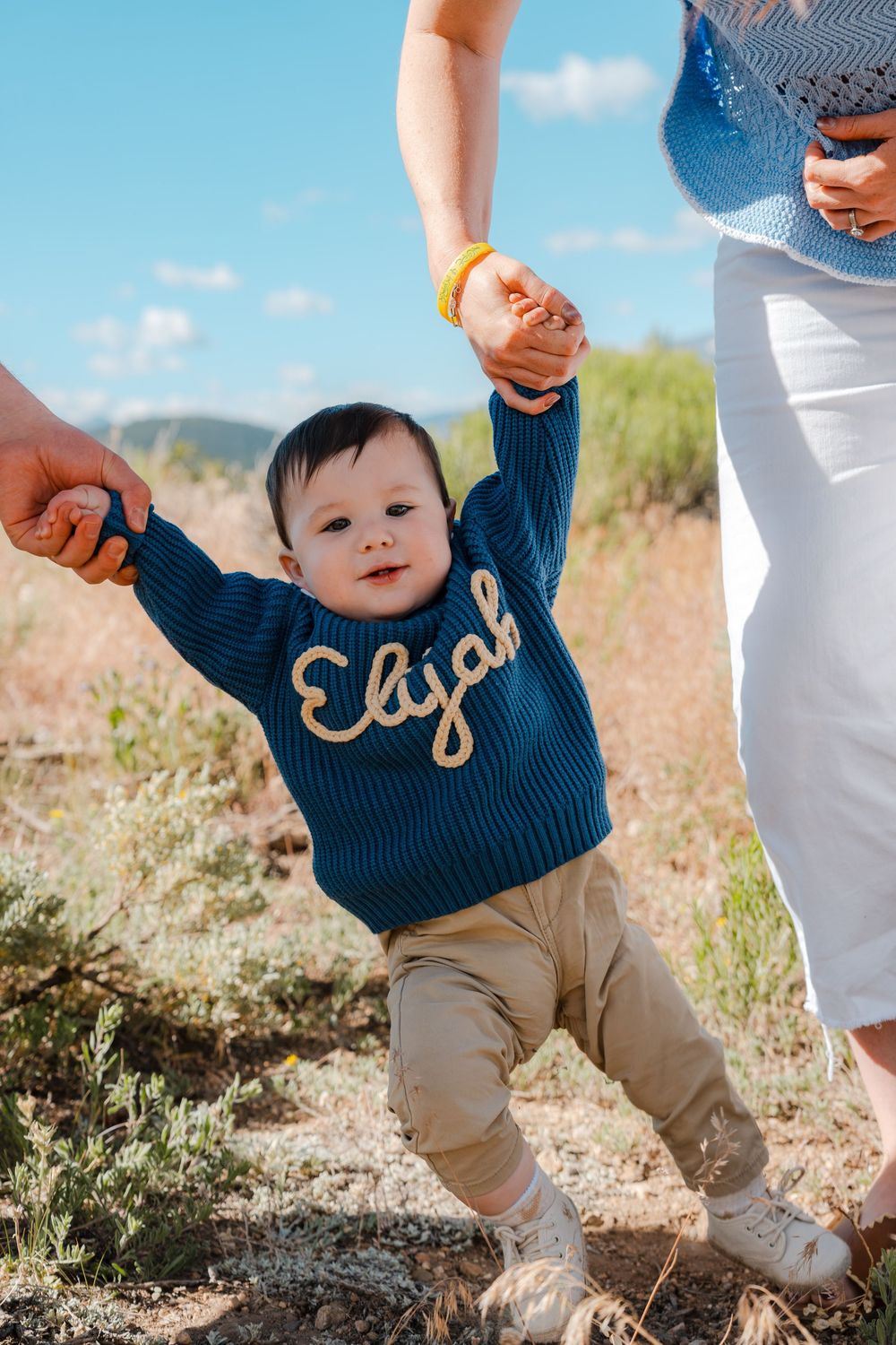 Baby Elijah in a teal knit sweater learning to walk outdoors, held by parents hands on a sunny day.