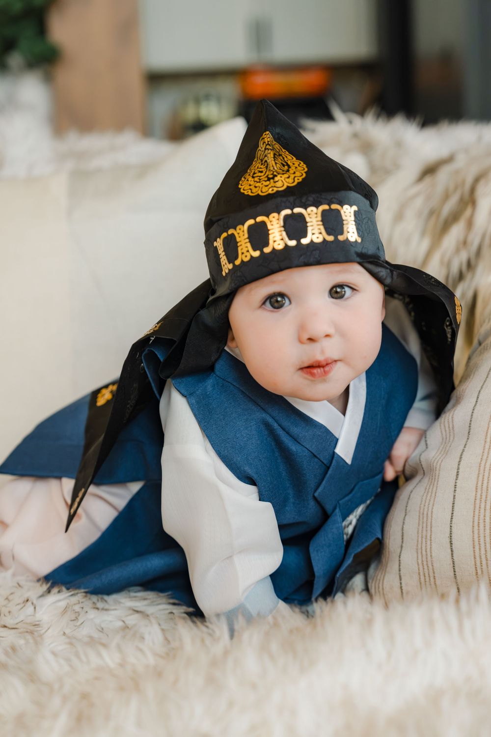 Adorable baby wearing traditional Korean hanbok outfit with decorative gat hat, resting on fluffy white cushions.