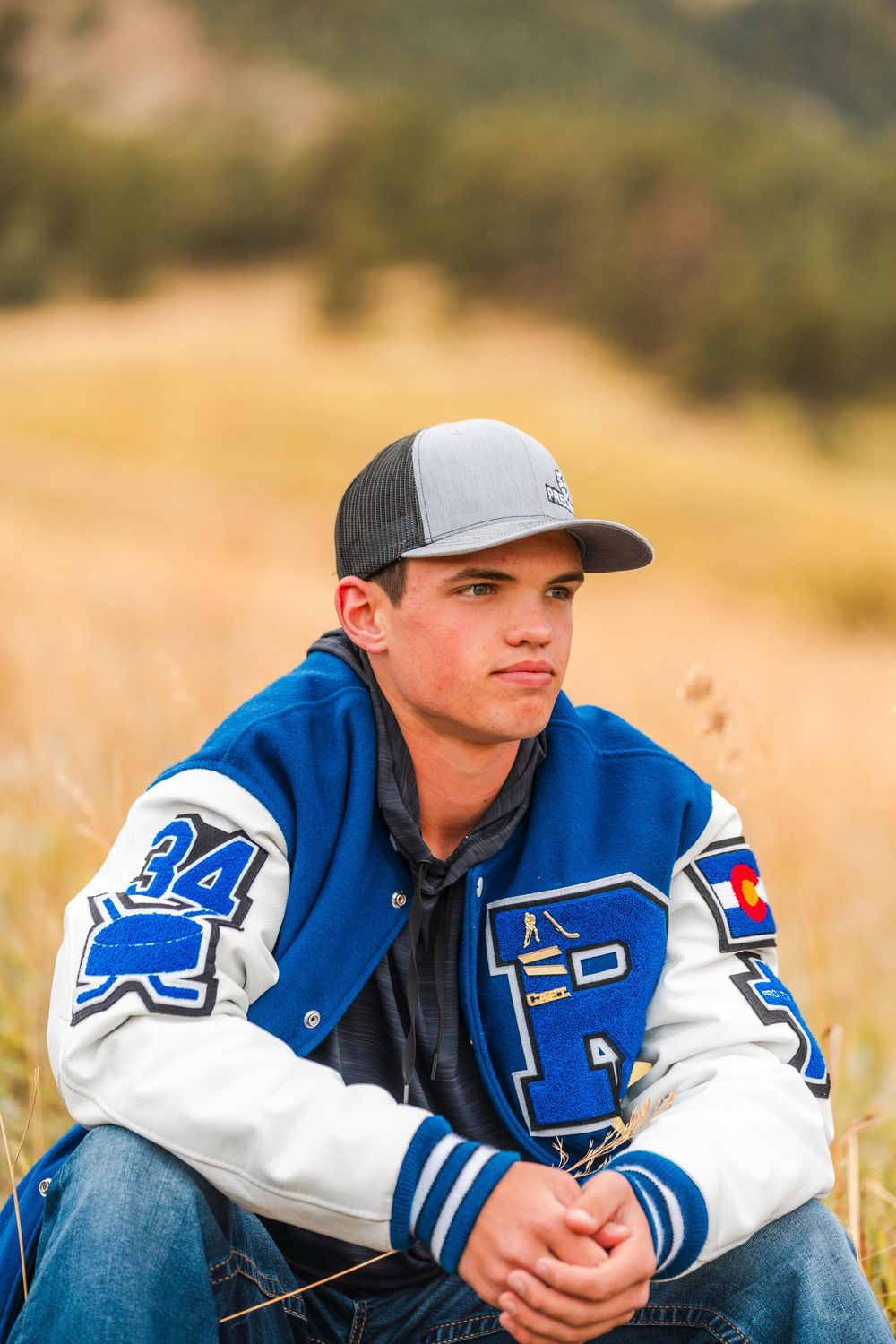 Young man in blue varsity letterman jacket and cap sitting in golden field outdoors.