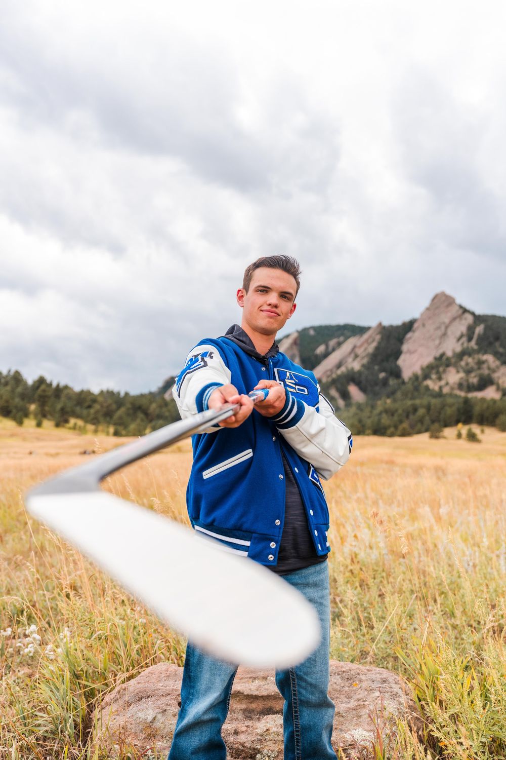 Teen in blue varsity jacket tossing baseball bat in mountain meadow under cloudy sky.