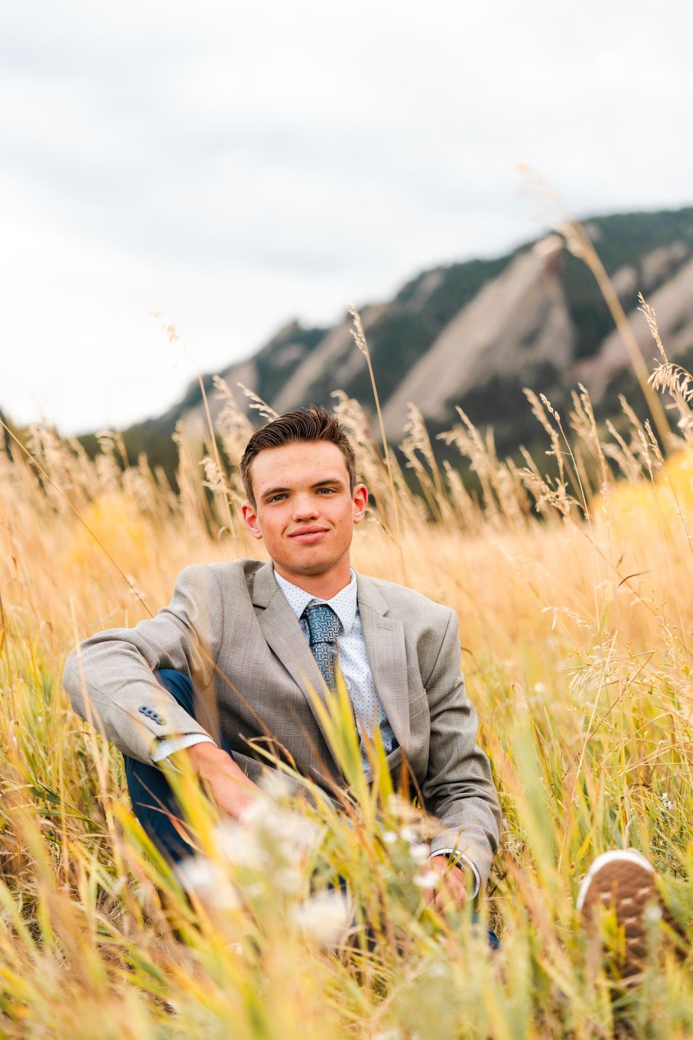 Teen male in gray blazer and tie sitting in tall grass with mountain flatirons backdrop.