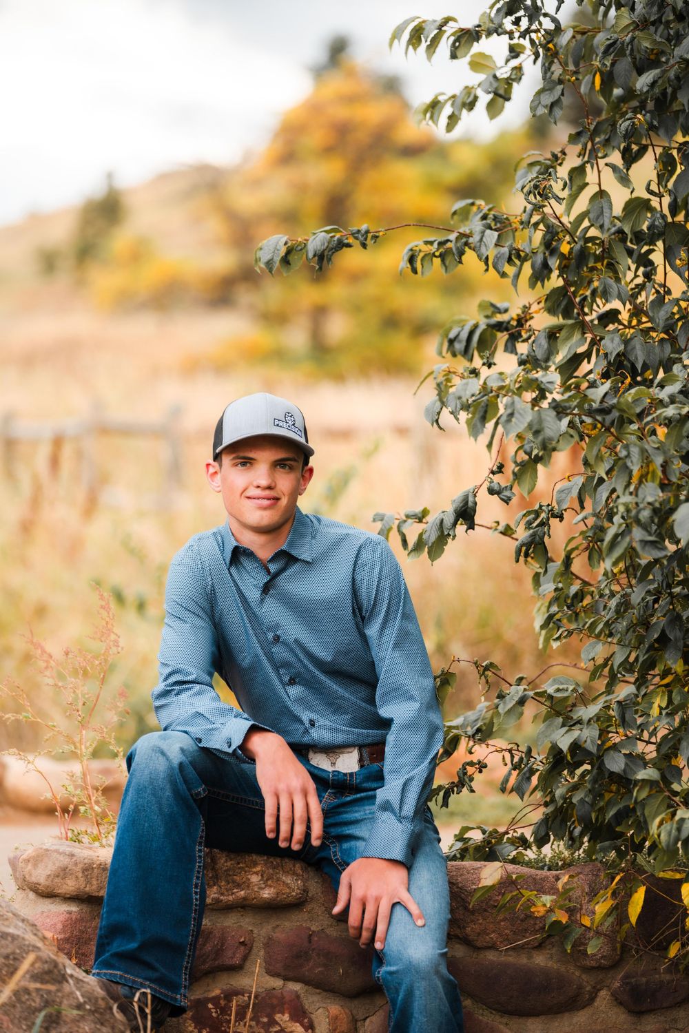 Young man in blue shirt and cap sitting on stone wall beside green foliage in fall.
