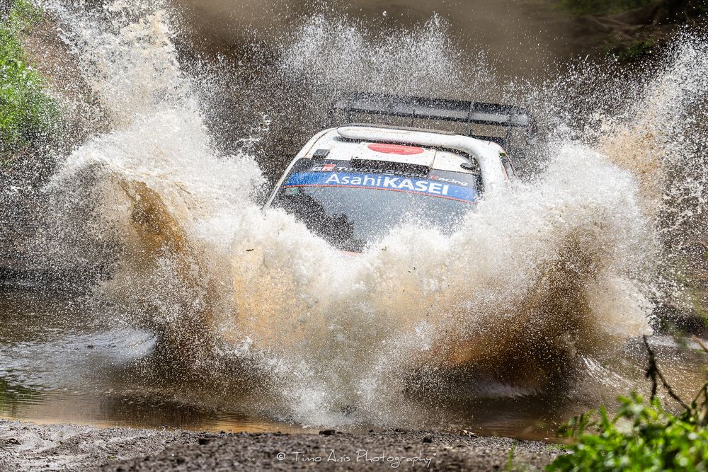 Rally car splashes through deep water creating dramatic spray during off-road race competition.