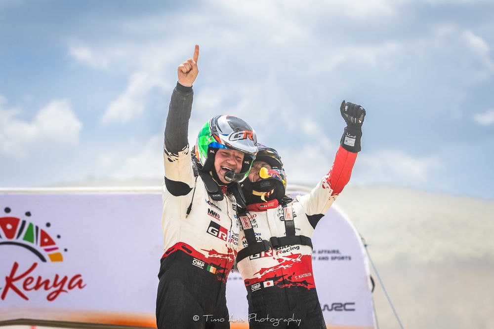 Rally car driver celebrates victory by raising fist in air while standing on top of vehicle at Kenya race event.