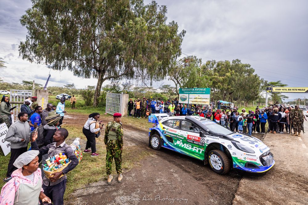 Rally spectators gather along a dirt road to watch race cars speed past during a competitive motorsport event.