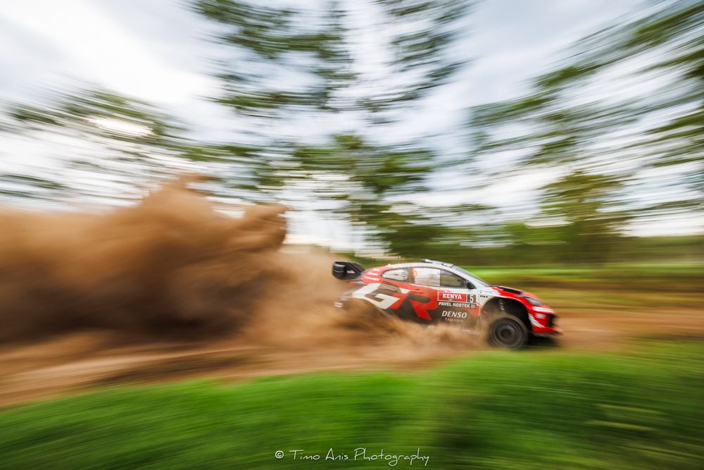Motion blur racing sequence of a red rally car speeding through a wooded course during competition.