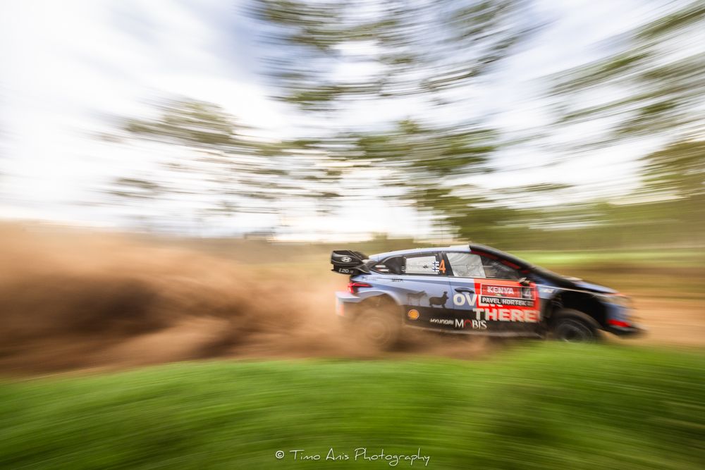 Rally car races through dirt track with motion blur effect creating dynamic speed impression during competition.