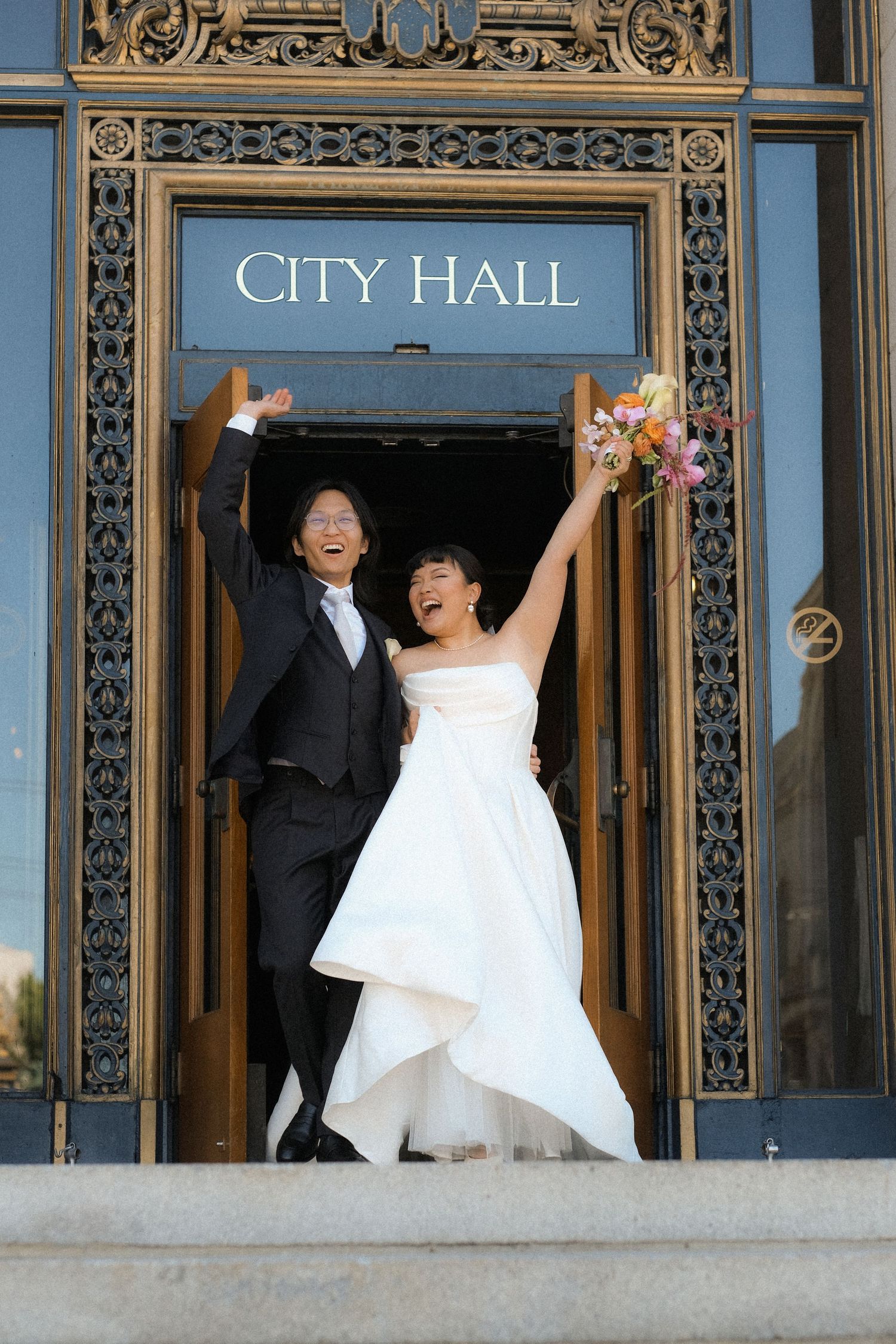 Joyful newlyweds celebrate while exiting City Hall with raised arms and flowers.
