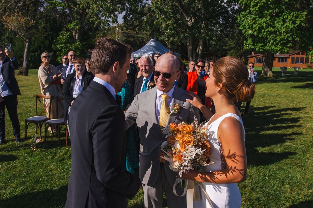 A wedding party gathers outdoors, with the bride and groom in the center holding a bouquet of flowers.