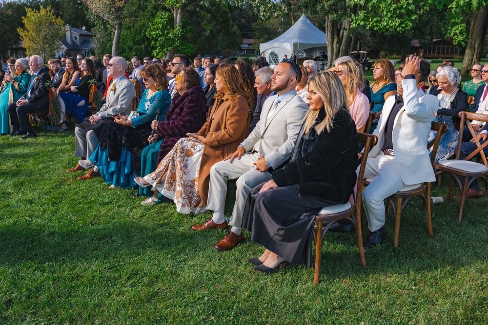 A group of people sitting in chairs on a grassy lawn, surrounded by trees and tents, attending an outdoor event.