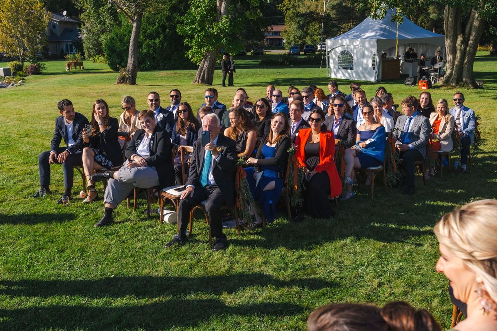 A large group of people sitting on the grass in a park, with tents and other outdoor equipment visible in the background.