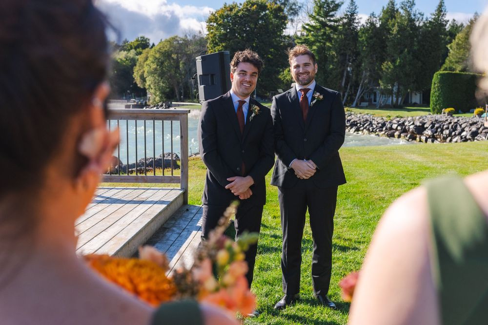 Two men in suits stand together outdoors in a grassy area with trees and a building in the background.