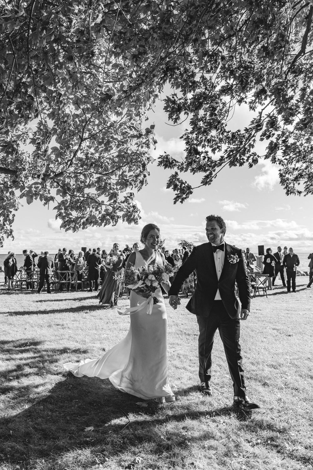 A black and white photograph of a bride and groom walking together on a grassy field surrounded by trees and guests.
